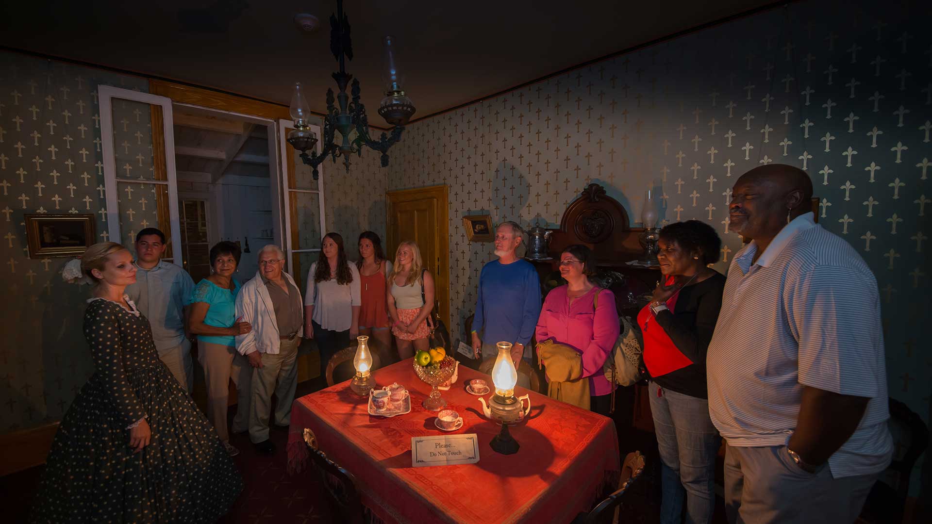 Guests and female ghost host in period costume standing in a dark room inside the Whaley House with a table in the middle that has two lanterns, a fruit bowl and plates and teacups