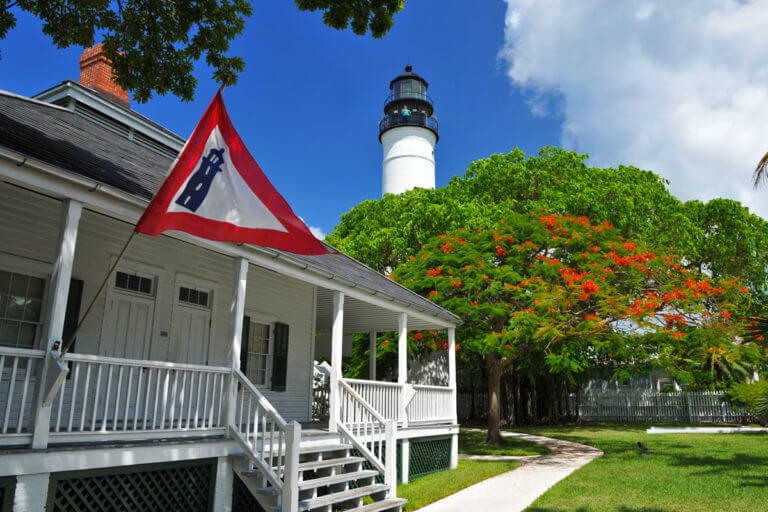 picture of old wooden house with stairs, porch, railing and windows, a flag with an icon of lighthouse in the foreground, and trees and the top of key west lighthouse in the background