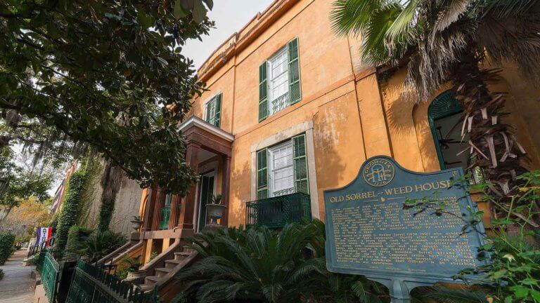 angle view of building exterior of sorrel weed house in savannah, georgia featuring two stone staircases leading to the entrance with shutter windows on either side and an iron sign to the right that reads 'Old Sorrel-Weed House'